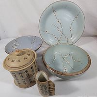 Front view of the five item lot showing three ceramic plates in light blue with floral branch design, cream colored script canister with lid, and small matching vase with textured handle and script.