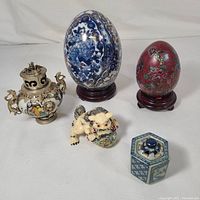Full view of the lot showing all five items including two decorative eggs on wooden stands, ceramic lion figurine, hexagonal ceramic container, and brass incense burner.