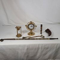 Full view of the vintage home decor items including brass clock, pipes, lacquered piece, hourglass candle holder, and decorative cane on white cloth background