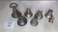 Overview photo of all eight antique bells showing variety of sizes, materials (brass and iron), and handle styles on plain tabletop.
