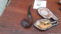 Overview photo showing both pipes on wooden surface, including the meerschaum pipe inside its fitted case and the dark wooden pipe beside it.