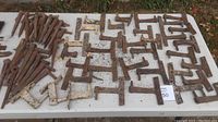 Wide view of a table covered with numerous hand forged iron shutter hardware pieces, mostly hinges and brackets. Some pieces have rust and white paint remnants.