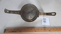 Full view of brass surveyor's compass set beside a ruler showing about 12 inches length. The compass dial is circular with an arm extending from each side for leveling.