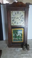 Full frontal view of wooden case wall clock with hand painted dial and landscape reverse glass tablet, weights positioned beside base.