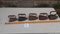 Wide shot showing all five sad irons lined up in size order on a white table with measuring stick