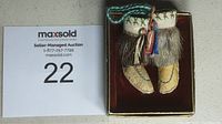 Pair of tiny Inuit doll boots on a dark fabric base inside a small box with a MaxSold auction card showing lot number 22.