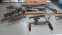 Wide view of various vintage hand tools spread on a flat surface including a metal shear, screwdrivers, combination square, spoke shaves, folding ruler, and other tools