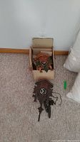 Two vintage wooden cuckoo clocks, one stored in a cardboard box, both on a carpeted floor.