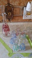 View of the glass decanter and thirteen apperitif glasses on a countertop showing red diamond pattern on decanter and varying blue/green patterns on glasses.