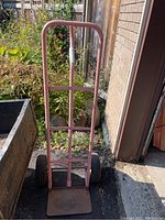 Front view of red metal framed dolly with two wheels, showing vertical handle and base plate, placed outdoors near a garden bed and garage entrance.
