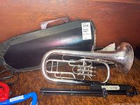 Silver euphonium with three valves placed in front of a black hard case and a folded black metal music stand on brown carpet.