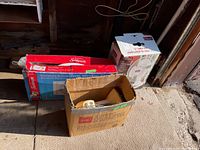 Three cardboard boxes on garage floor; Classic 12-inch table fan box, Sunbeam twin window fan box, and a box with disassembled Electrohome fan parts.