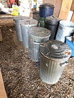Four metal garbage cans arranged in a row on gravel under a lean-to, showing worn and rusted lids and handles