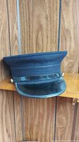 Blue fabric uniform cap with a black shiny visor and button sides, shown at an angle on a wood shelf against a wood paneled wall.