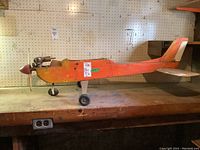 Full side view of the wooden model plane body showing the orange paint, mounted gas engine, propeller spinner, and tail assembly without wings.