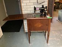 Full view of Singer sewing machine mounted in wood cabinet with fold-out table extension and door on the front of the cabinet.