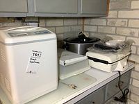 White Black and Decker bread maker, Crockpot slow cooker, white Hamilton Beach grill, and white cake carrier on basement countertop next to sink.