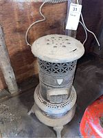 Full view of the antique metal stove showing the top perforated cover, cylindrical body, and ornate legs. Dust is visible on the surface.
