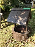 Wooden wishing well painted dark brown with an octagonal slatted base and a pitched roof; placed outdoors on grass with surrounding greenery.