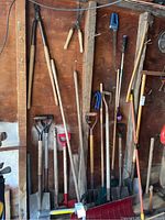 Photo of multiple yard tools including shovels, rakes, and hoes leaning against a wooden wall