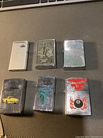 Photo showing six assorted lighters on a surface, including two Zippo style metal lighters with distinct designs.