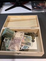 Open wooden box containing mixed old coins and paper currency including 500 Indonesian Rupiah and 20 Israeli Shekel bills.