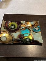 Top-down view of rectangular enamel tray with raised edges and multicolor circular pattern placed on dark wood table.