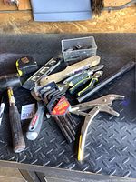 Photo showing assortment of hand tools including locking pliers, spirit levels, tape measure, hammer, and utility knives arranged on a black diamond plate surface.