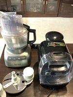 KitchenAid food processor with clear processing bowl, control panel, and dicing kit attachments on table alongside black Tassimo coffee machine.