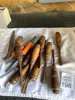 Lot of wood chisels with wooden handles and a couple of orange-handled chisels laid out on a white towel, along with metal files including a rasp.