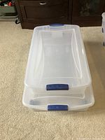 Two clear plastic under bed storage containers stacked, showing blue locking handles and white lids.