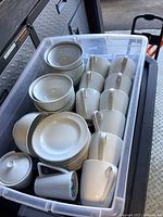 A plastic container filled with white porcelain cups with handles, side plates, and the creamer and sugar bowl with lid.