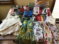 Overview of all items in lot showing large assortment of aprons, towels, napkins, pot holders, oven mitts, and tablecloths arranged on a table.