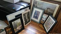 Framed art pieces leaning against wall on wooden floor, varying sizes and visual styles.