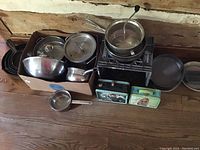 Overview of cast iron skillets, mixing bowls, Maitre Chef pots, lunchboxes, and pie pans on wooden floor
