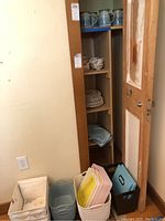 View of shelving with ceramic tankards on middle shelves and storage bins on floor, including plastic and cloth bins in multiple colors and sizes.