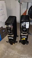 Two black Honeywell electric heaters with metal fins, on caster wheels, standing on a concrete floor near shelving.