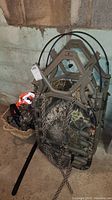 Side view of folded camouflage ladder stand tree seat with attached safety straps and harness, along with orange reflective vests and camouflage clothing on floor basket.