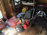 Front-left perspective of red steel cutting deck, engine, and handlebars inside a shed