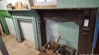 Wide shot showing three fireplace mantles side by side against basement wall; dark brown carved mantle on right and two green painted mantles on left