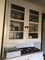 White wooden cabinet with glass doors containing picture frames, ceramic wall hangings shaped like birds and crosses, a retro sign, and small mirrors