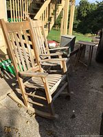 Two weathered wooden rocking chairs with slatted backs and seats, positioned outdoors on a concrete surface with natural sunlight.