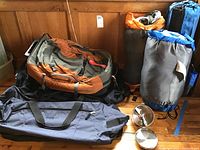 Photo shows two sleeping bags in carrying bags, a campfire pot with lid and utensils, a large backpack, two duffel bags, and a carry-on bag arranged on a wooden floor.