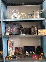 Shelf with various vintage kitchenware, decor items, toys and books including a Season's Greetings sign, collection of toy cars, ceramic teapot, silver pitcher, and baseball glove.