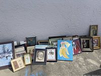 Photo showing a row of framed artwork including portraits, landscapes, and abstract pieces, along with several empty frames, arranged outdoors against a wall.