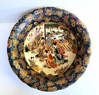 Top view of large blue rimmed porcelain bowl with a central painted scene of Japanese geisha women in traditional kimonos and a garden setting. Rim decorated with gold floral and butterfly designs.