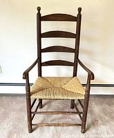 Front view of antique ladderback armchair showing four curved horizontal slats, ball finials, woven rush seat, and wooden frame with visible wear.