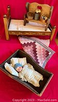 Antique Armand Marseille baby doll laid in a dark wooden cradle with cloth pillows and quilt, wooden cradle and bench with accessories on display.