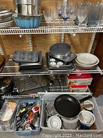 Wide view of metal bakeware, glassware, muffin pans, frying pans, and two boxes of Ball canning jars, along with bins of assorted kitchen utensils and cookware.