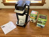 Front-side image of black Keurig K15 coffee maker, two boxes of Green Mountain Coffee pods, and instruction manual on wooden table.